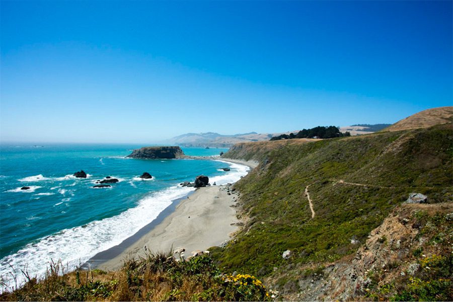 On the coast of the Pacific Ocean: blue ocean with white crashing waves on a small beach with green hills. Some rocks in the distance in the water.