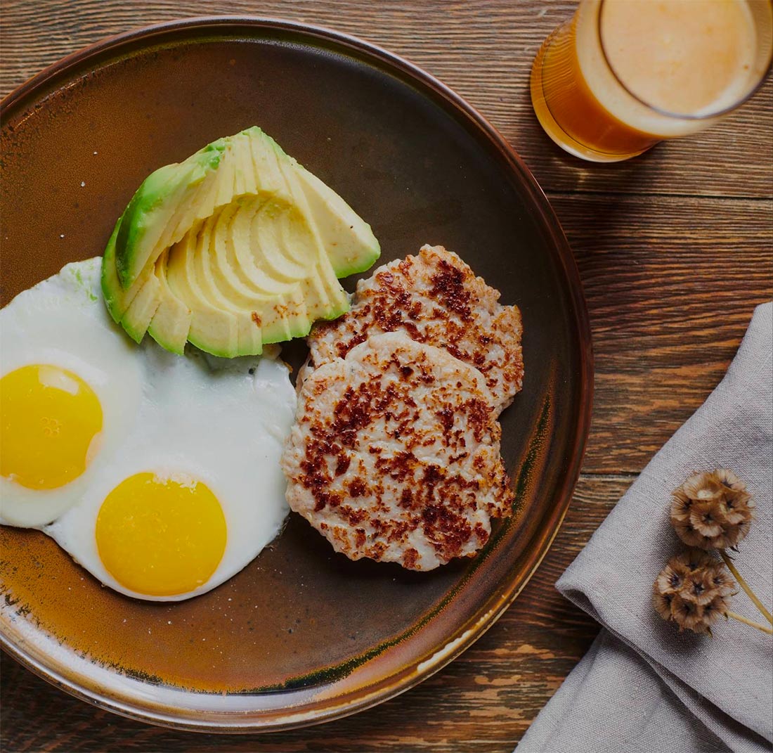 Overhead view of a breakfast plate with two eggs over easy, sausage patty and fresh sliced avocado along with a glass of juice.