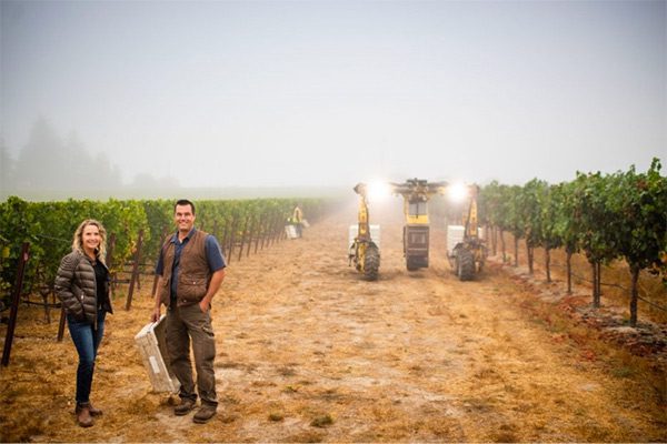 on the vineyard, man and woman smile for photo with grape trees and farming machinery in the background.