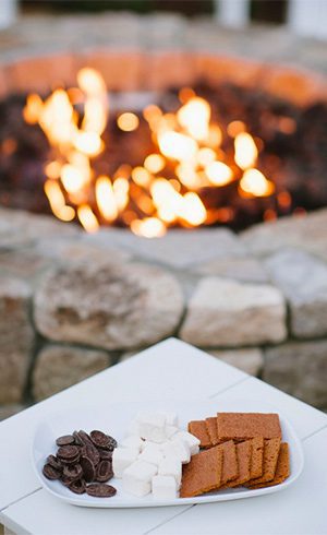 close-up of s'mores on a white plate with a stone fire-pit with fire lit in the background.