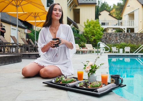 Young woman in swimming attire next to Farmhouse Inn pool with a coffee cup and a tray with two place settings with plates of salads, juice and flowers.