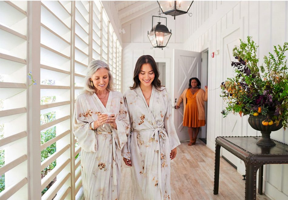 An older woman and younger woman walking happily in matching robes from the Farmhouse Inn Spa. The Spa keeper is in the background.