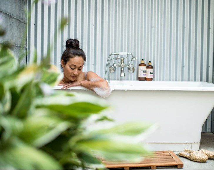 white bathtub with woman in it with plant greenery in the foreground.