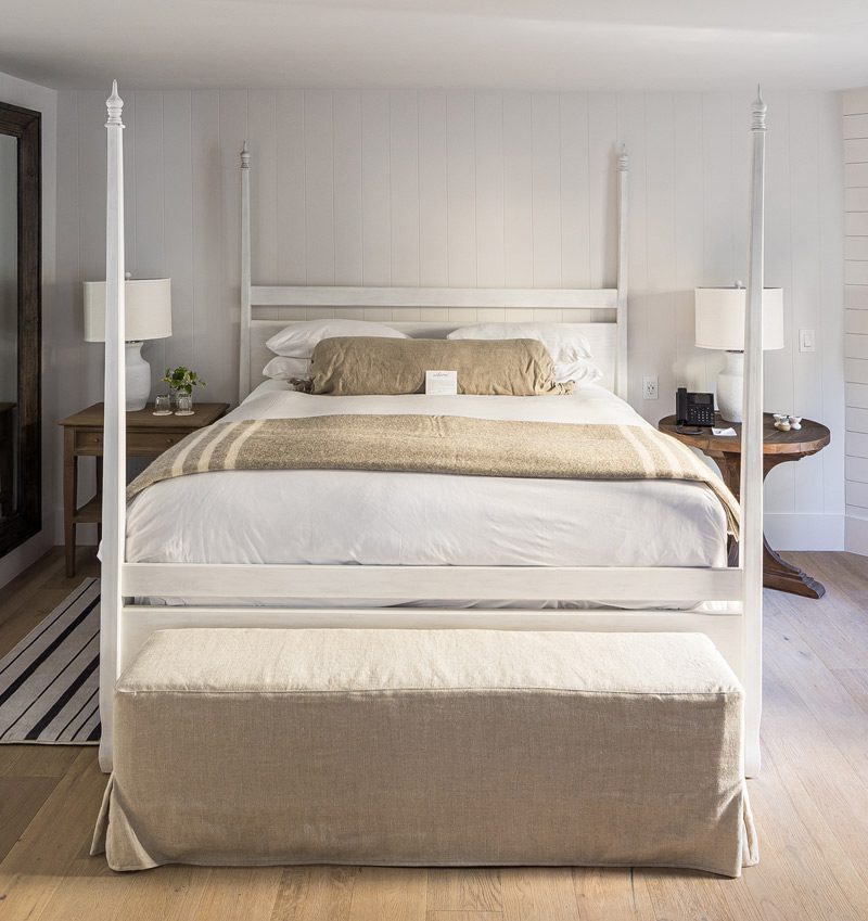 Interior view of Cottage Petite Suite facing bed. Large bed with white bed posts and light bedding, sideview of fireplace with fire, white cushioned chair, and large wall mirror with wood frame. Large paneled wood floors with striped area rug next to bed.