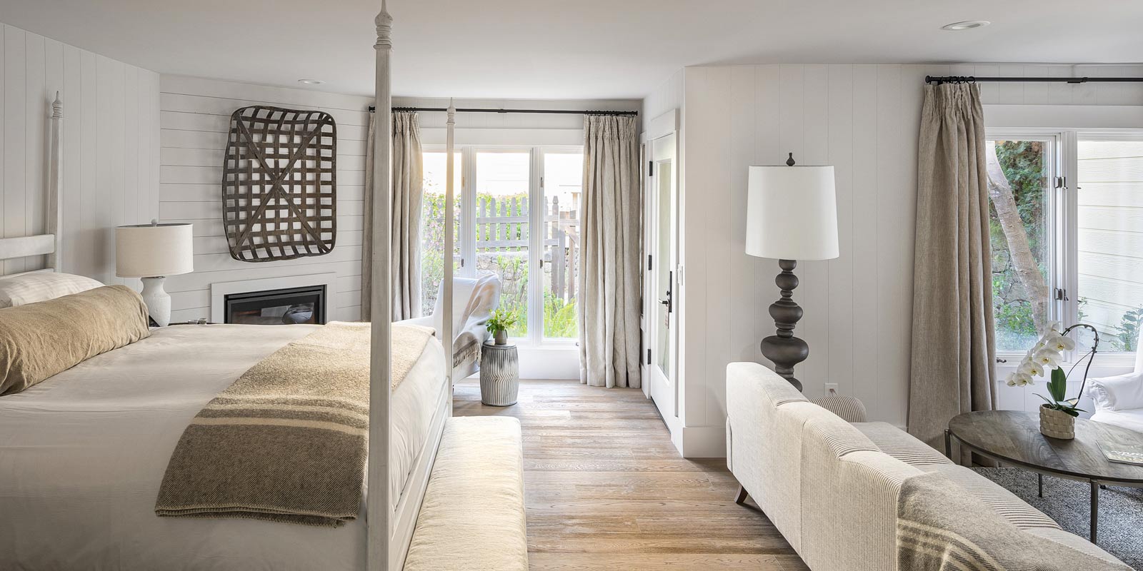 Interior view of Cottage Petite Suite. Foot of large bed with white bed posts and light bedding, sideview of light-colored couch, large elegant floor lamp, wall decorations, two glass doors in back of room.