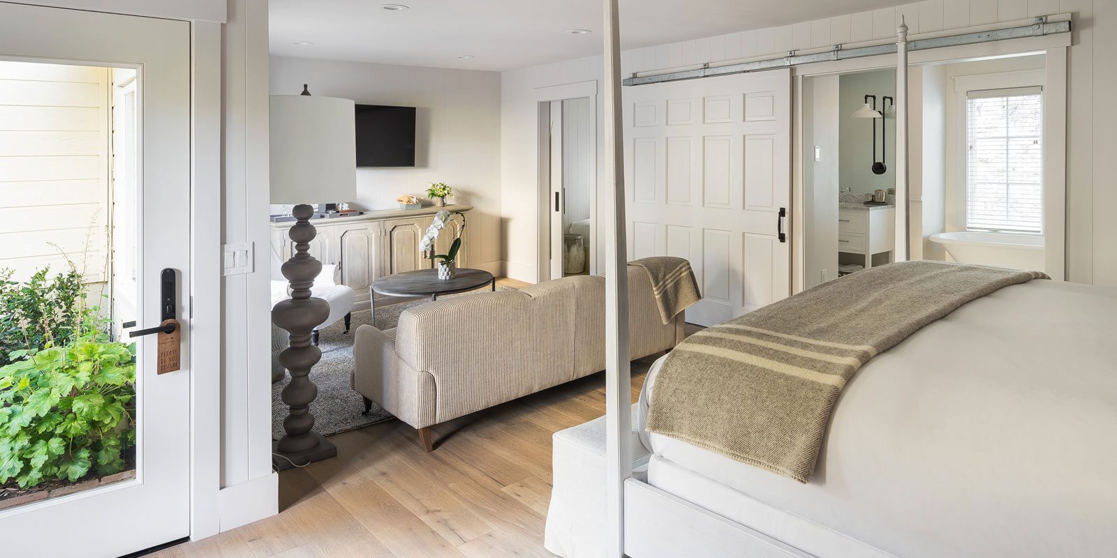 Interior view of Cottage Petite Suite. Foot of large bed with white bed posts and light bedding, back of light love seat, open sliding door with glimpse of bathroom and tub, glass door.