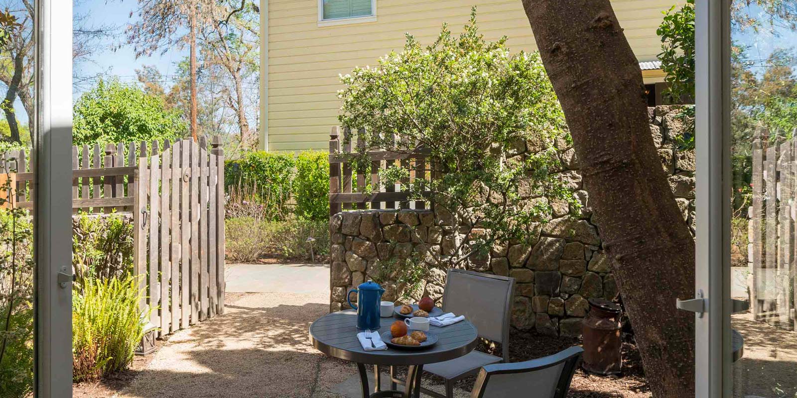 Patio outside Cottage Petite Suite with small round table and two patio chairs with two place settings. Wood picket fence in background and quaint yellow two floor dwelling.