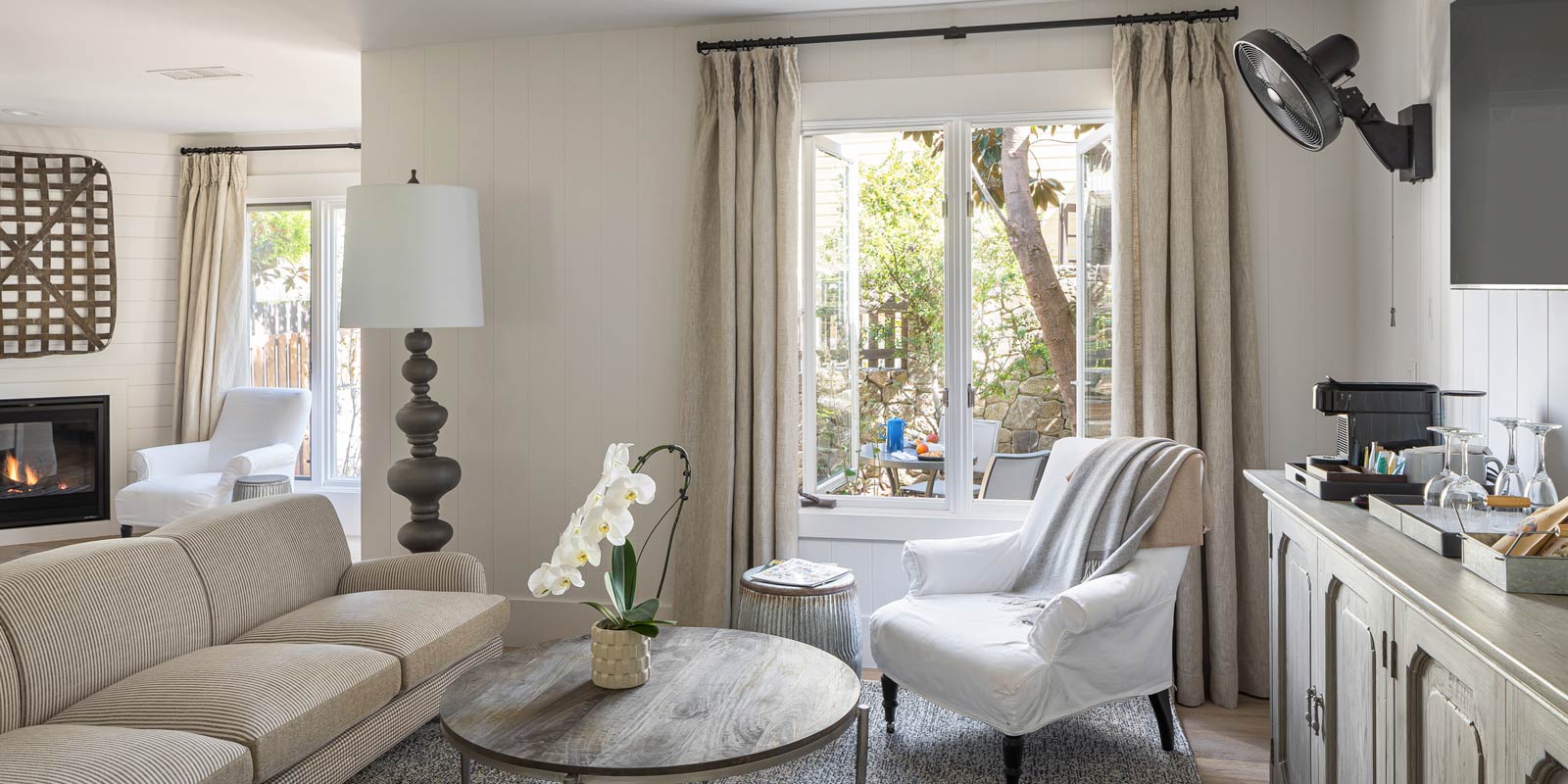 Interior view of Cottage Petite Suite facing large window. Side view of couch wood round coffee table, white cushioned chair, area rug, credenza, mid sized flat screen-mounted, large fancy floor lamp, fireplace with fire on.