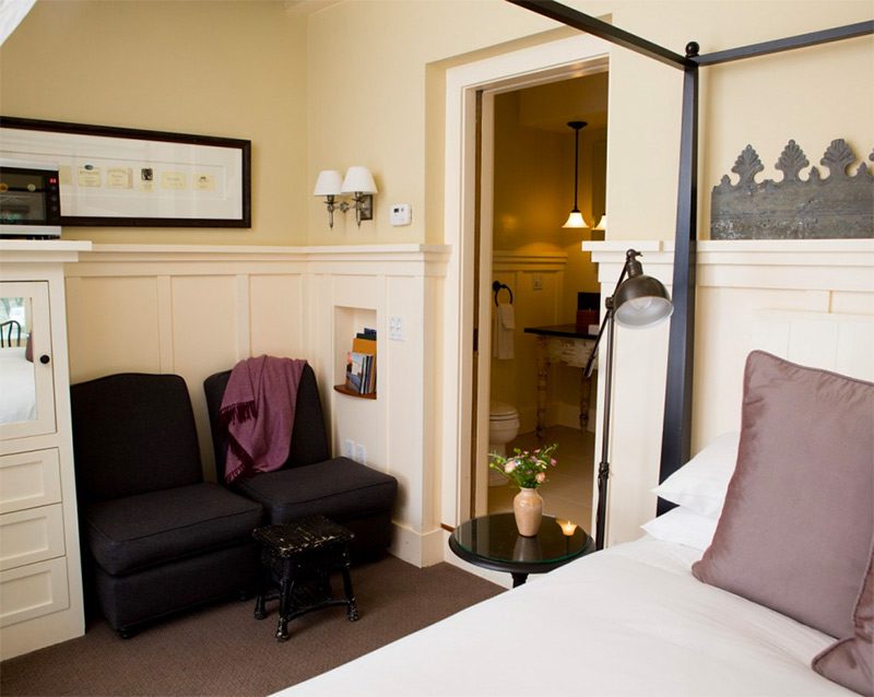 Farmhouse Room Interior: Two black cushioned chairs, portion of bed with white bedding and small black round nightstand with flowers and lit candle, open door into bathroom