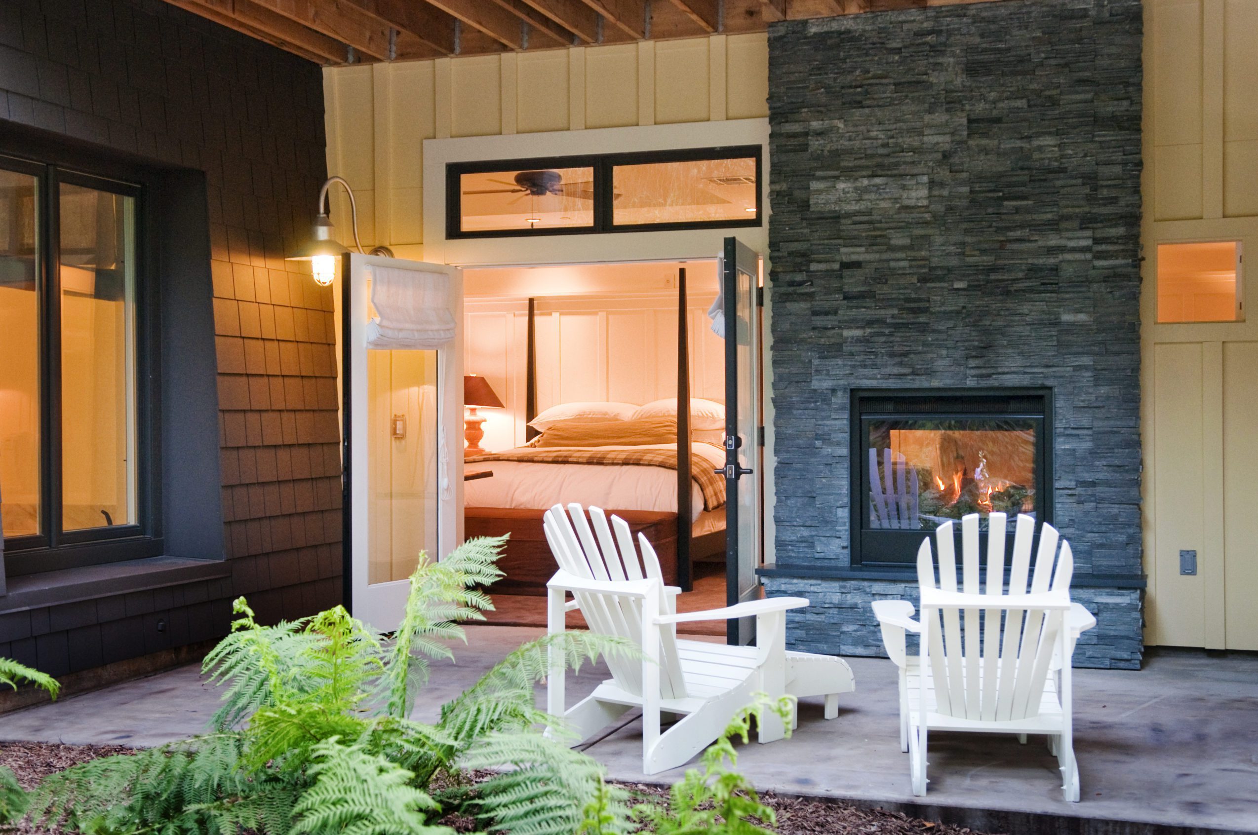 porch view of Barn Junior Suite featuring indoor, outdoor fireplace, white patio wood chairs, and a view inside room with large bed with white bedding