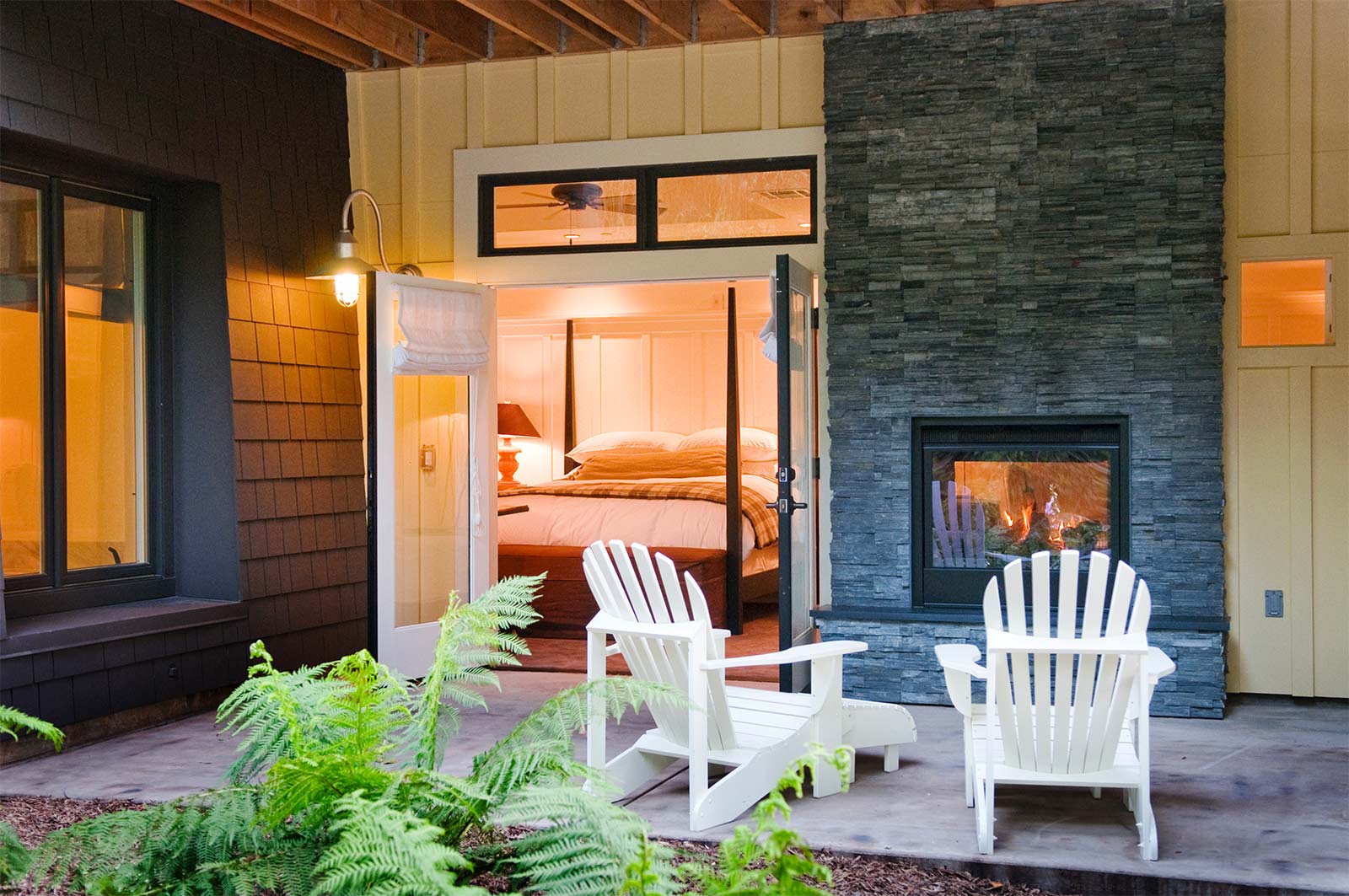 porch view of Barn Junior Suite m featuring indoor, outdoor fireplace, white patio wood chairs, and a view inside room with large bed with white bedding