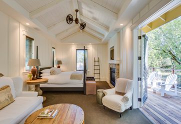 interior of Barn Junior Suite with ceiling fan, large bed with white bedding, white chair and love seat. porch door is open to a wood deck with white patio chairs