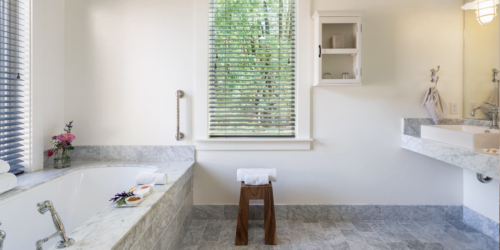 interior of large, elegant bathroom: gray tiled bathtub, matching tiled floors, large vertical glass window, elegant sink and mirror to the right. looking out to greenery,