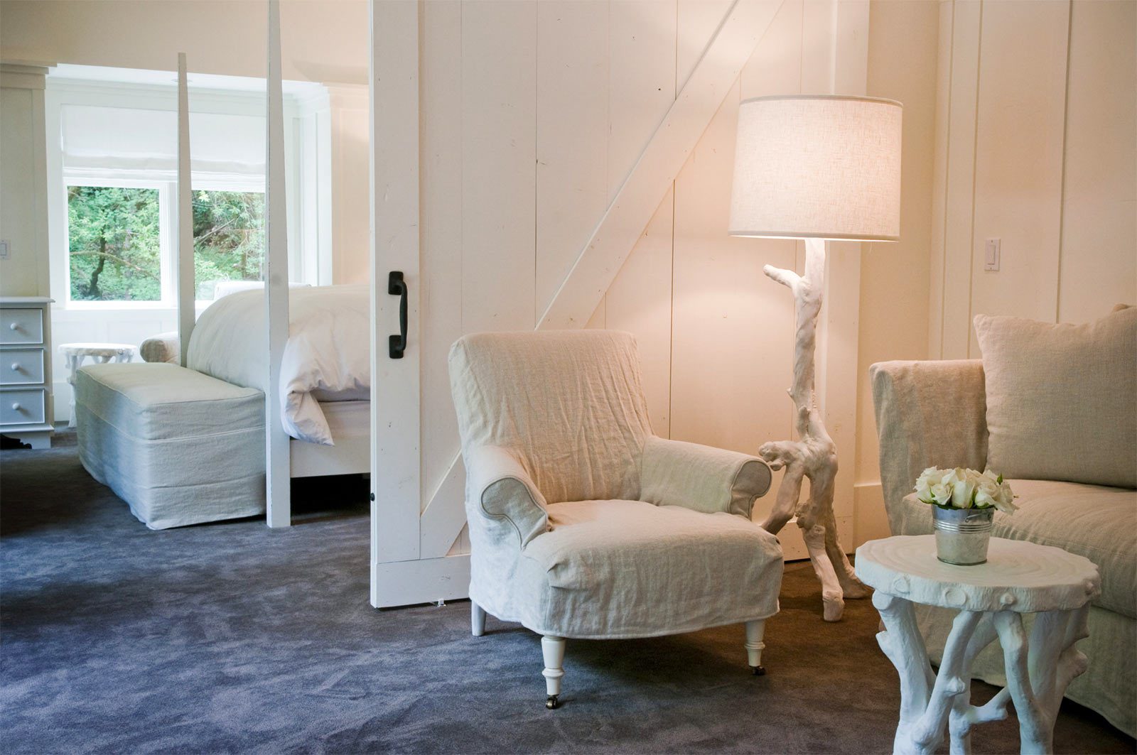 Interior of Barn One Bedroom Suite: Dark blue carpet, white cushioned chair and love seat. foot if large bed with white bedding in background with sliding glass door.