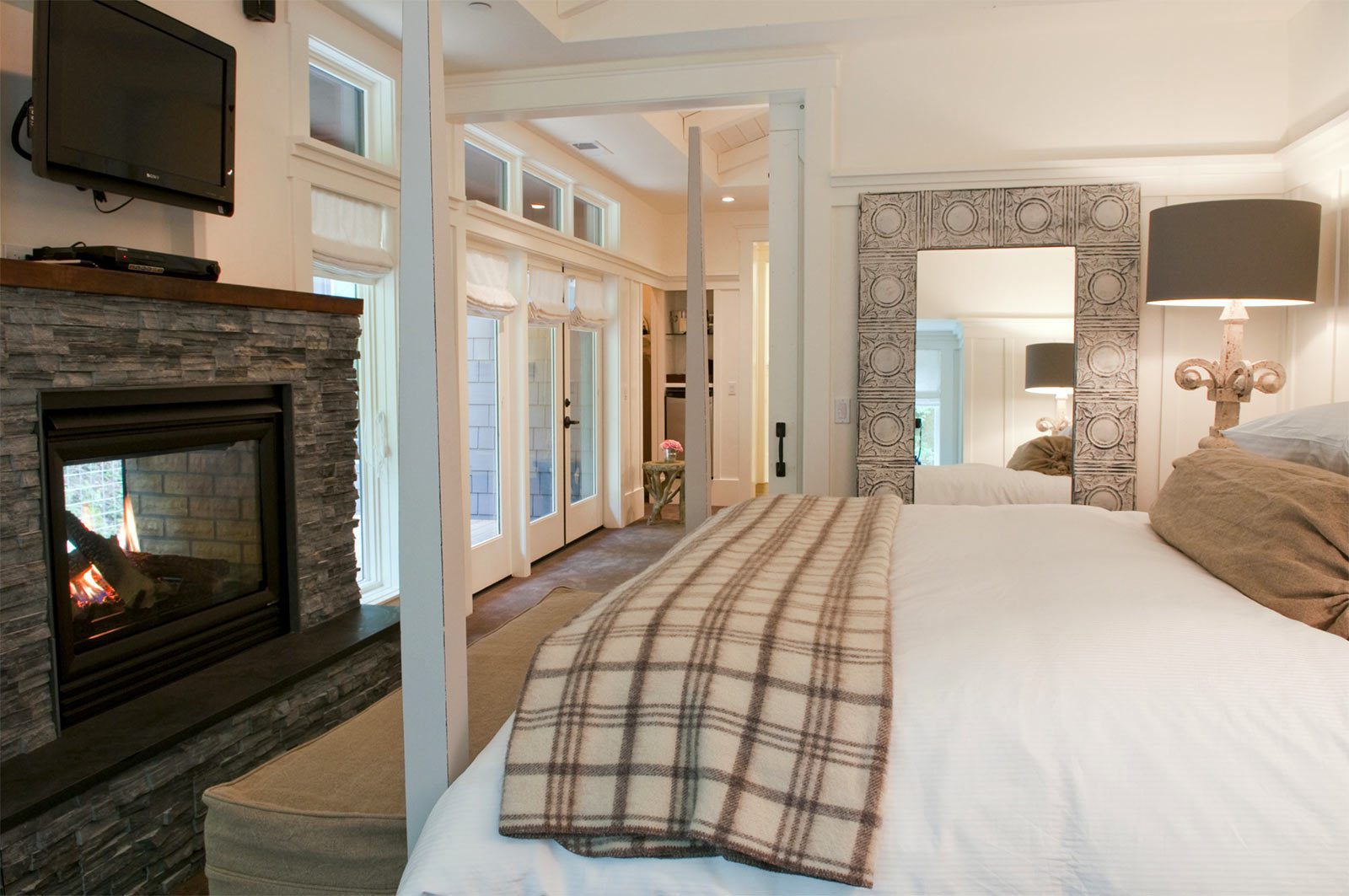 Interior of Barn One Bedroom Suite: side view of large bed with white bedding and plaid folded blanket at foot of bed. On the left is a dark stone gas fireplace with flatscreen TV over it. There are large floor to ceiling windows on the wall.