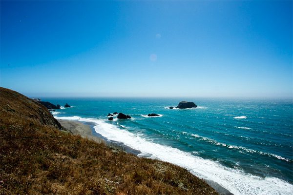 On the coast of the Pacific Ocean: blue ocean with white crashing waves on a small beach with green hills. Some rocks in the distance in the water.
