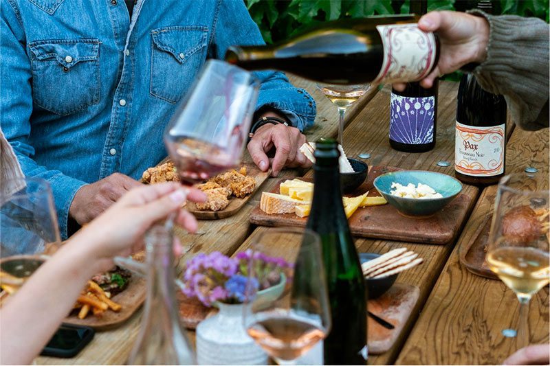 Outdoor gathering people sitting at a wood table with various food items and a man pouring Freeman wine.