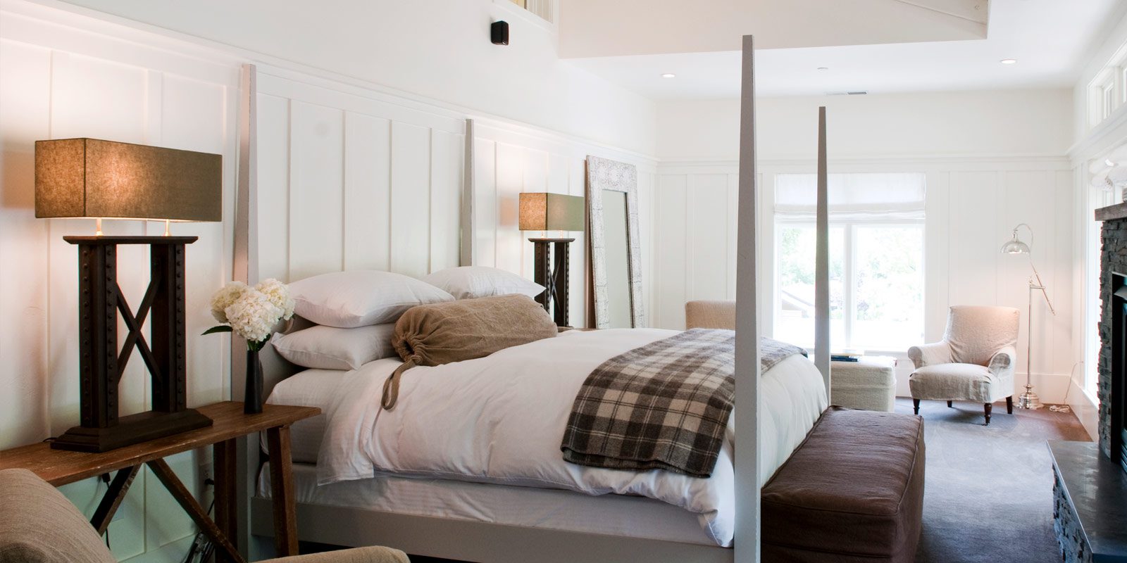 Interior view of Barn Junior Suite. White walls, stone fireplace with mounted TV above, large bed with white bedding with gray bedposts, high ceilings with ceiling fan.