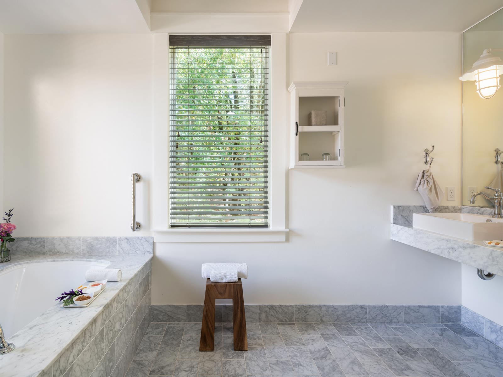 interior of large, elegant bathroom: gray tiled bathtub, matching tiled floors, large vertical glass window, elegant sink and mirror to the right. looking out to greenery,