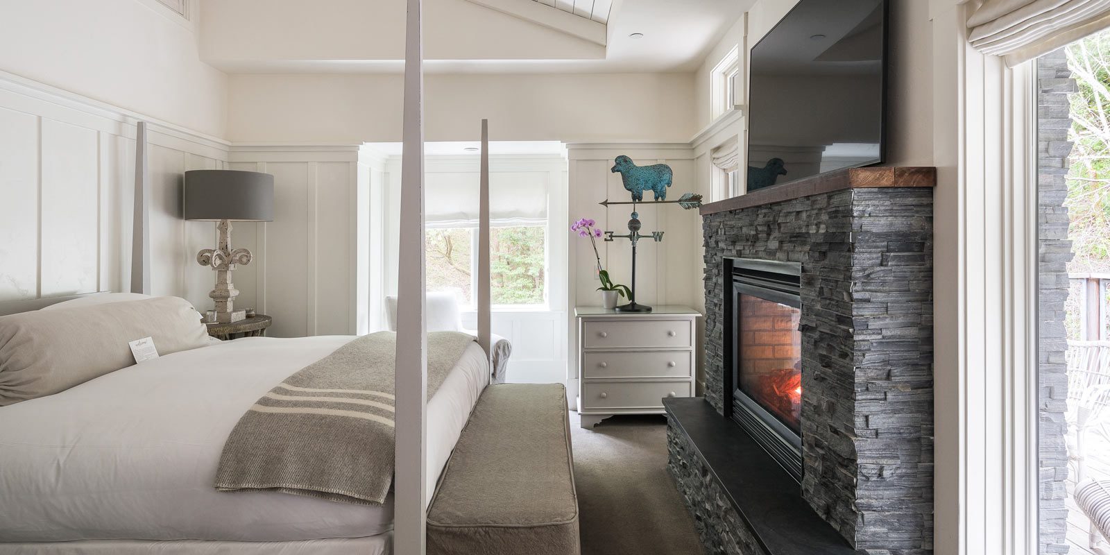 Interior of Barn One Bedroom Suite: side view of large bed with white bedding and plaid folded blanket at foot of bed. On the right is a dark stone gas fireplace with flatscreen TV over it. There is a large glass door on the back wall.