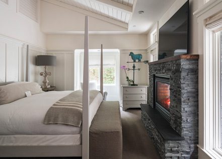 Interior of Barn One Bedroom Suite: side view of large bed with white bedding and plaid folded blanket at foot of bed. On the right is a dark stone gas fireplace with flatscreen TV over it. There is a large glass door on the back wall.