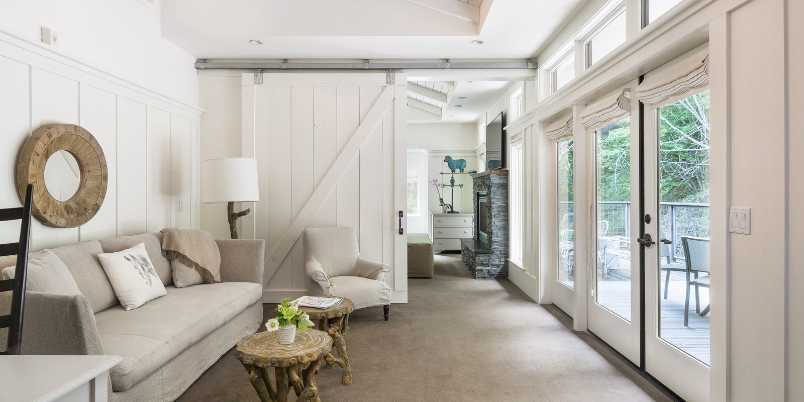 Interior of Barn One Bedroom Suite: side view of large bed with white bedding and plaid folded blanket at foot of bed. On the left is a nice sitting area with beige couch and on the right is large glass doors leading to the wood porch.