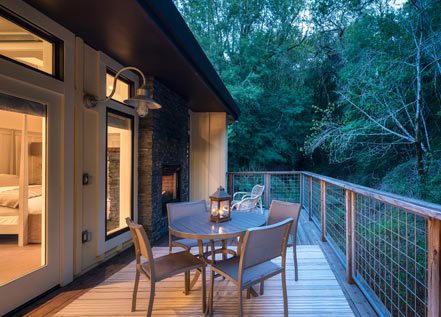 Elevated wood porch of Barn One Bedroom Suite with round patio table with chairs, wood bannister with greenery in back and glimpse into the room.