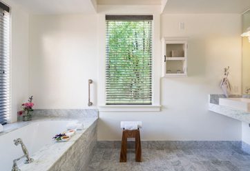 interior of large, elegant bathroom: gray tiled bathtub, matching tiled floors, large vertical glass window, elegant sink and mirror to the right. looking out to greenery,