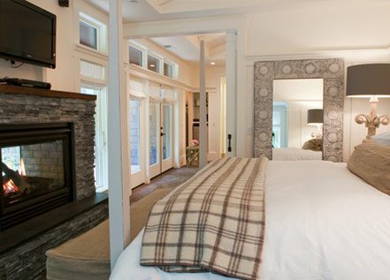 Interior of Barn One Bedroom Suite: side view of large bed with white bedding and plaid folded blanket at foot of bed. On the left is a dark stone gas fireplace with flatscreen TV over it. There are large floor to ceiling windows on the wall.