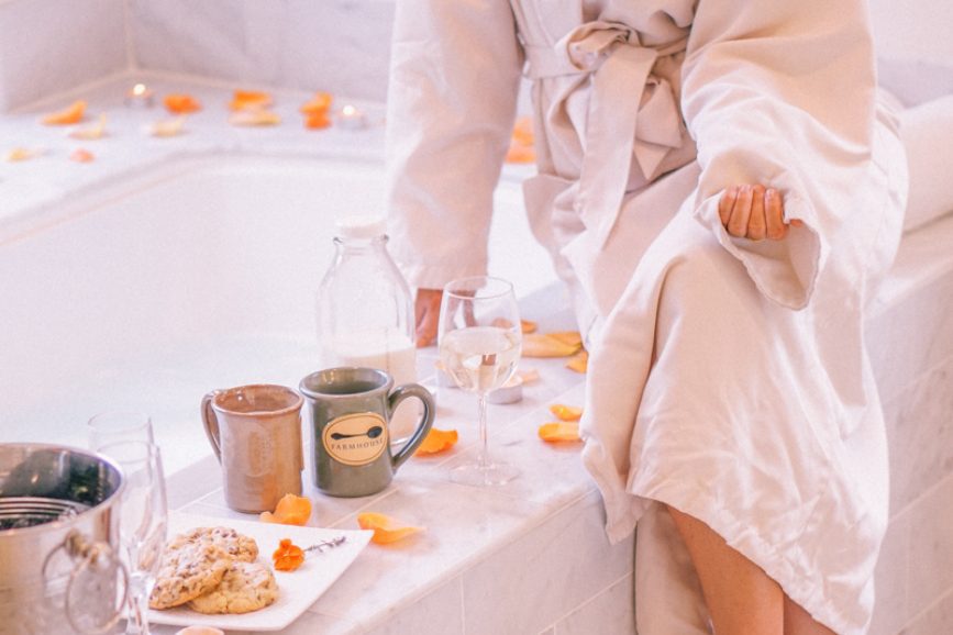 Farmhouse guest sitting on bathtub ledge, enjoying the Candlelight Tub Turndown special. Rose petals, tea, Farmhouse mug, and desserts line the tub