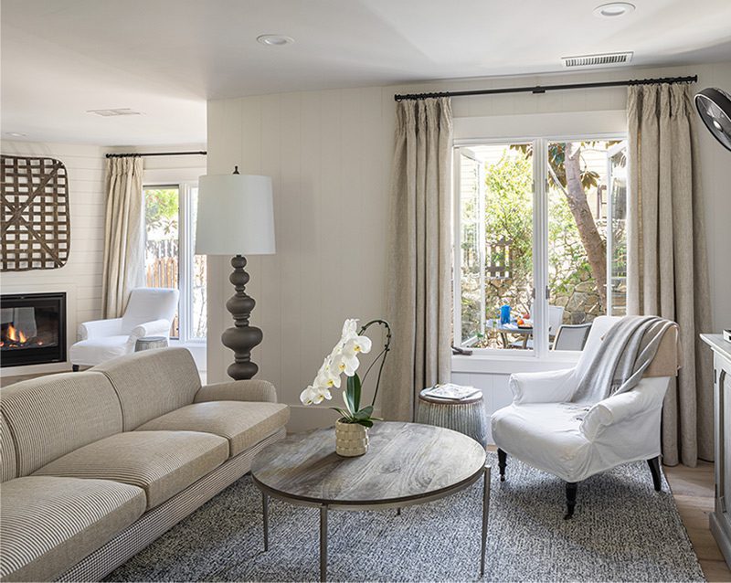 Interior view of Cottage Petite Suite facing large window. Side view of couch wood round coffee table, white cushioned chair, area rug, credenza, mid sized flat screen-mounted, large fancy floor lamp, fireplace with fire on.