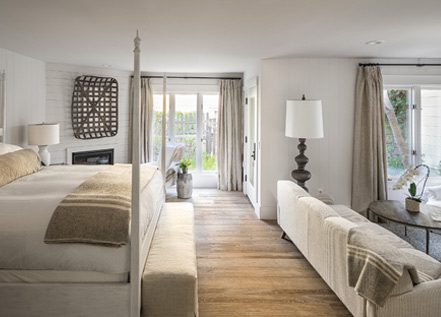 Interior view of Cottage Petite Suite. Foot of large bed with white bed posts and light bedding, sideview of light-colored couch, large elegant floor lamp, wall decorations, two glass doors in back of room.