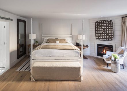 Interior view of Cottage Petite Suite facing bed. Large bed with white bed posts and light bedding, sideview of fireplace with fire, white cushioned chair, and large wall mirror with wood frame. Large paneled wood floors with striped area rug next to bed.