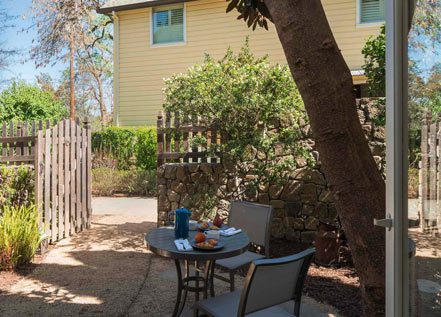 Patio outside Cottage Petite Suite with small round table and two patio chairs with two place settings. Wood picket fence in background and quaint yellow two floor dwelling.