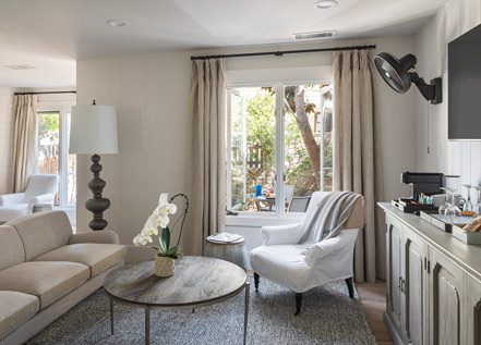 Interior view of Cottage Petite Suite facing large window. Side view of couch wood round coffee table, white cushioned chair, area rug, credenza, mid sized flat screen-mounted, large fancy floor lamp, fireplace with fire on.