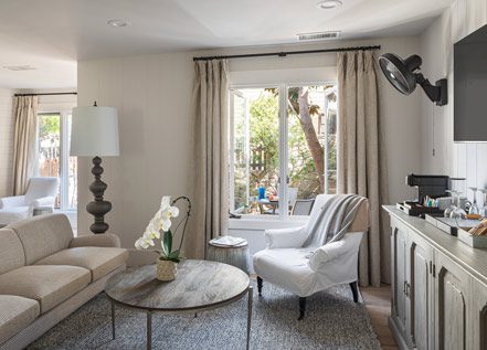 Interior view of Cottage Petite Suite facing large window. Side view of couch wood round coffee table, white cushioned chair, area rug, credenza, mid sized flat screen-mounted, large fancy floor lamp, fireplace with fire on.