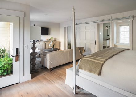 Interior view of Cottage Petite Suite. Foot of large bed with white bed posts and light bedding, back of light love seat, open sliding door with glimpse of bathroom and tub, glass door.