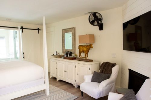 Interior of Cottage Room: side view of large bed with white bed posts and white bedding. Counter-height dresser on other side of room with lamp and mirror hung above.