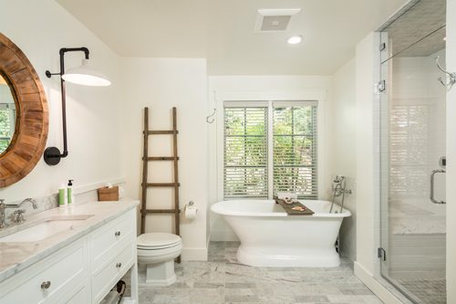 Interior of Cottage Room Bathroom: white walls, sink and counter to left with drawers and round mirror hung above, elegant stand-alone bathtub in back with vertical windows above, white toilet with ladder decoration on wall, glass door for shower on right.