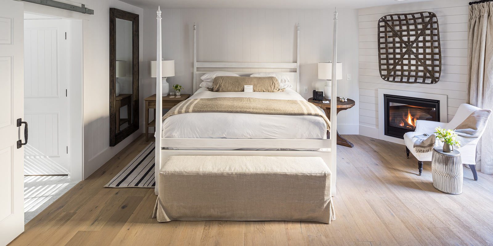 Interior view of Cottage Petite Suite facing bed. Large bed with white bed posts and light bedding, sideview of fireplace with fire, white cushioned chair, and large wall mirror with wood frame. Large paneled wood floors with striped area rug next to bed.