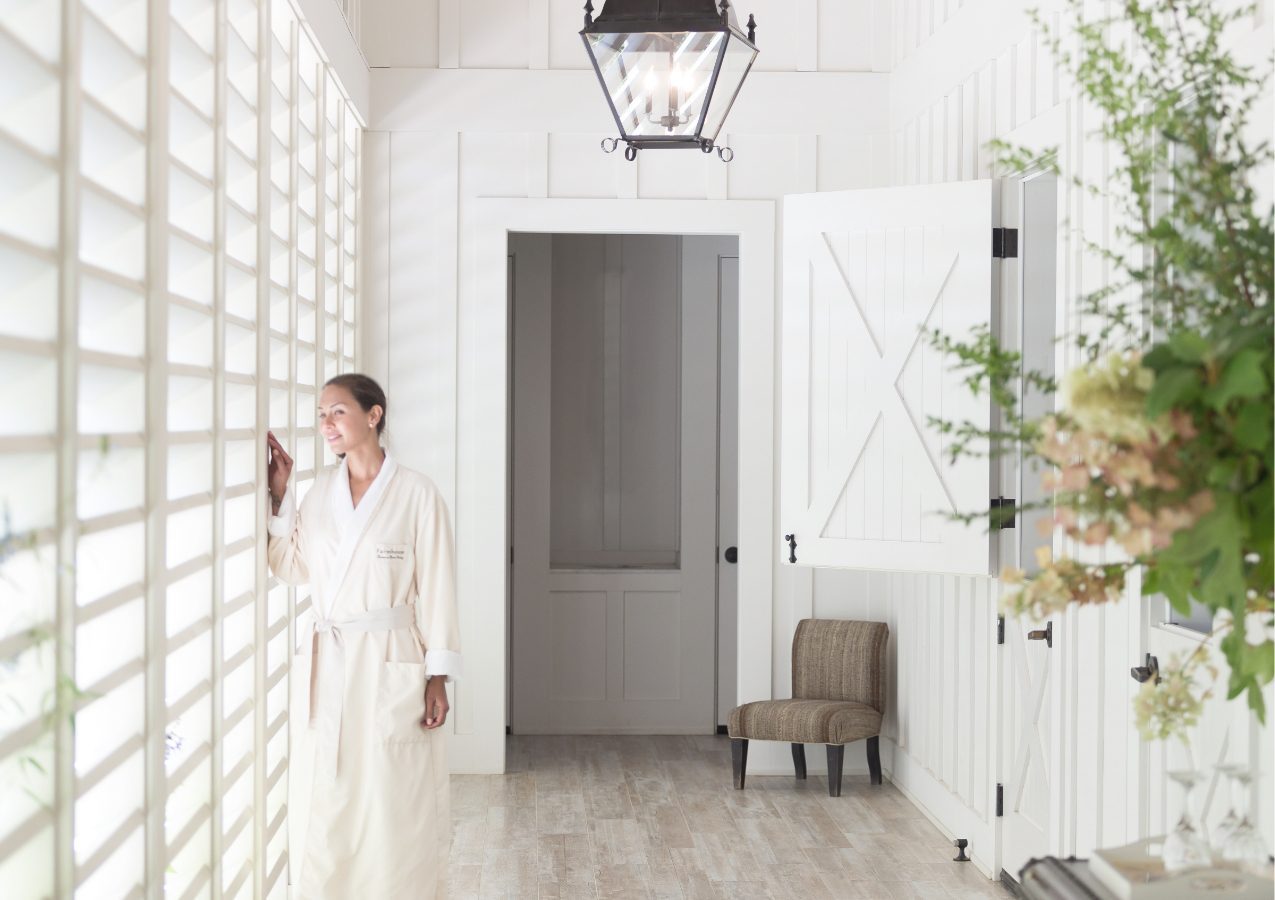 Hallway of the Farmhouse Spa. Windows with blinds on the left, wood floors, elegant lantern lights above and white wood walls. Woman peaking out of the blinds looking out into the Farmhouse property.