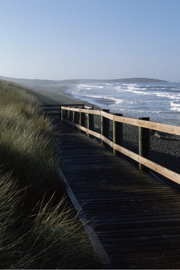 On the coast of the Pacific Ocean: blue ocean with white crashing waves on a small beach with green hills. Some rocks in the distance in the water.