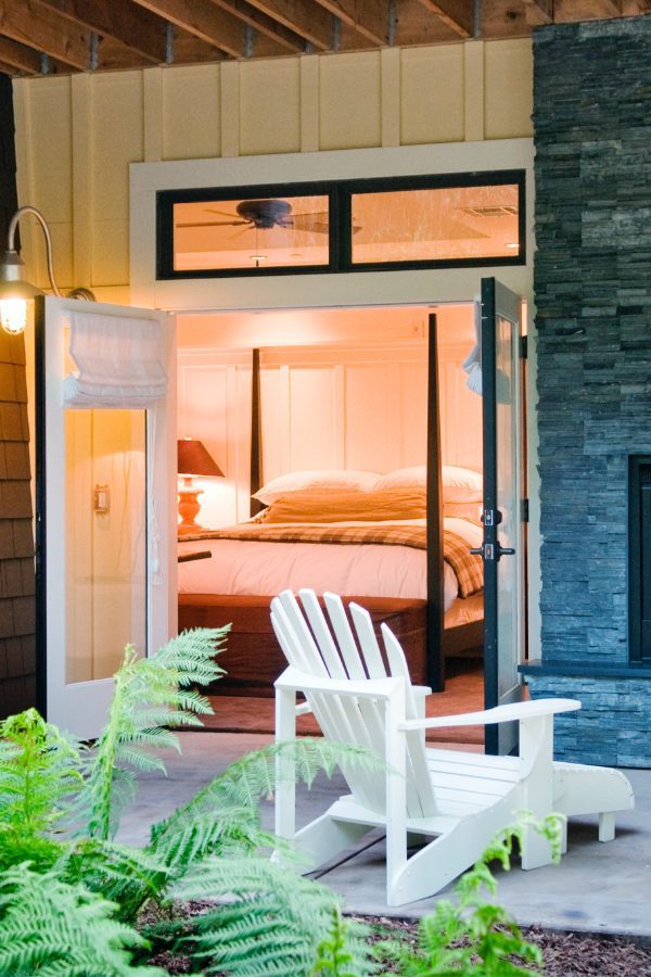 porch view of Barn Junior Suite featuring indoor, outdoor fireplace, white patio wood chairs, and a view inside room with large bed with white bedding