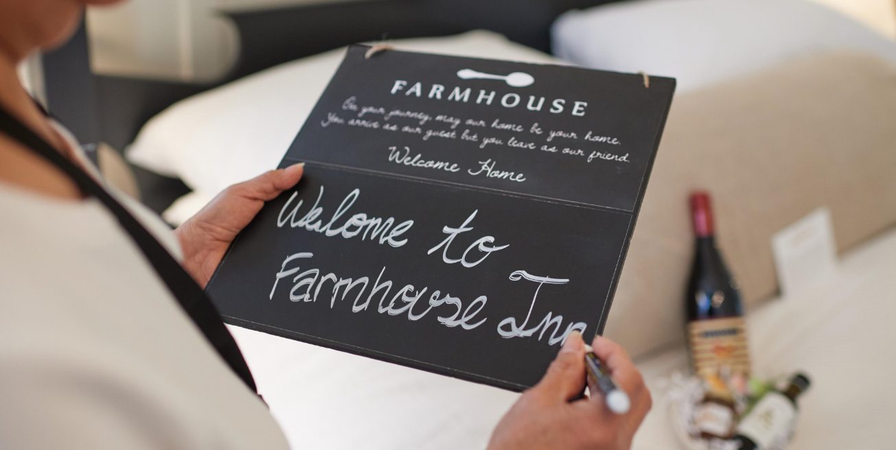 Farmhouse Inn employee preparing the welcome sign, wine and gift, on the bed in a room for a guest's arrival.