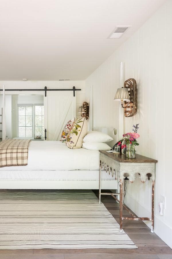 Interior of Cottage Room: side view of large bed with white bed posts and white bedding with plaid folded blanket at foot of bed. Counter-height dresser on other side of room with lamp and mirror hung above. Glass doors in back leading to patio with lush greenery.