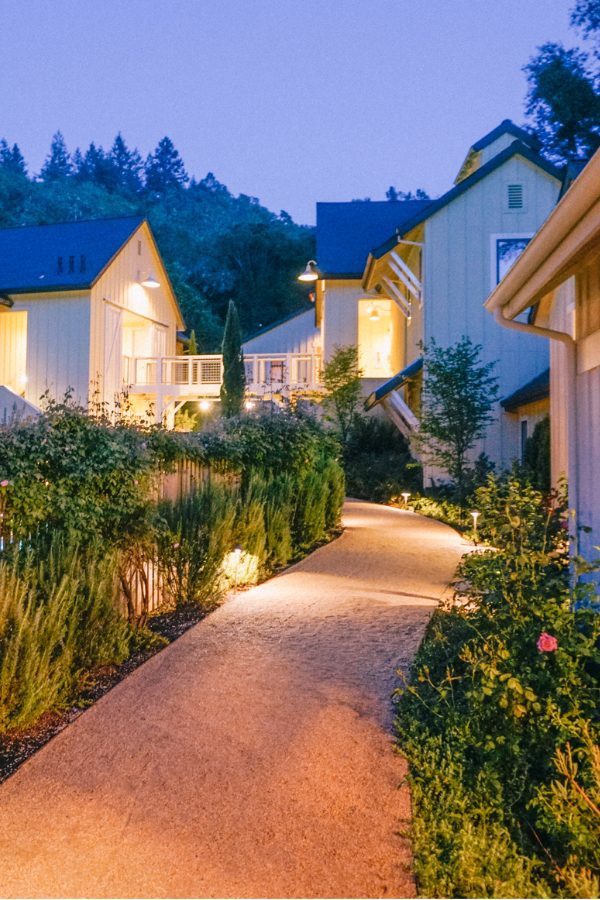 Quaint pathway on the perimeter of one of the Farmhouse Inn buildings surrounded by lush greenery and pink flowers at dusk. .