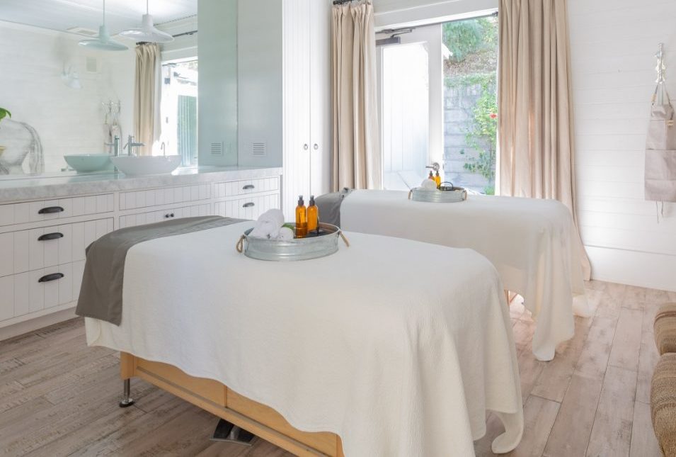Spa room with massage table with tray of massage products, white wood cabinetry with sink and full wall mirror, glass door with a view of a patio.
