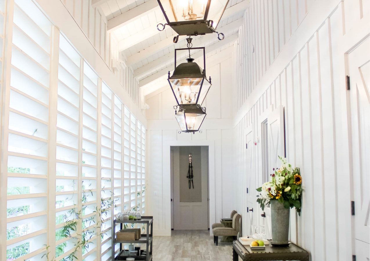 Hallway of the Farmhouse Spa. Windows with blinds on the left, wood floors, elegant lantern lights above and white wood walls.