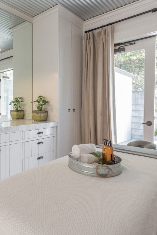 Spa room with massage table with tray of massage products, white wood cabinetry with sink and full wall mirror, glass door with a view of a patio.