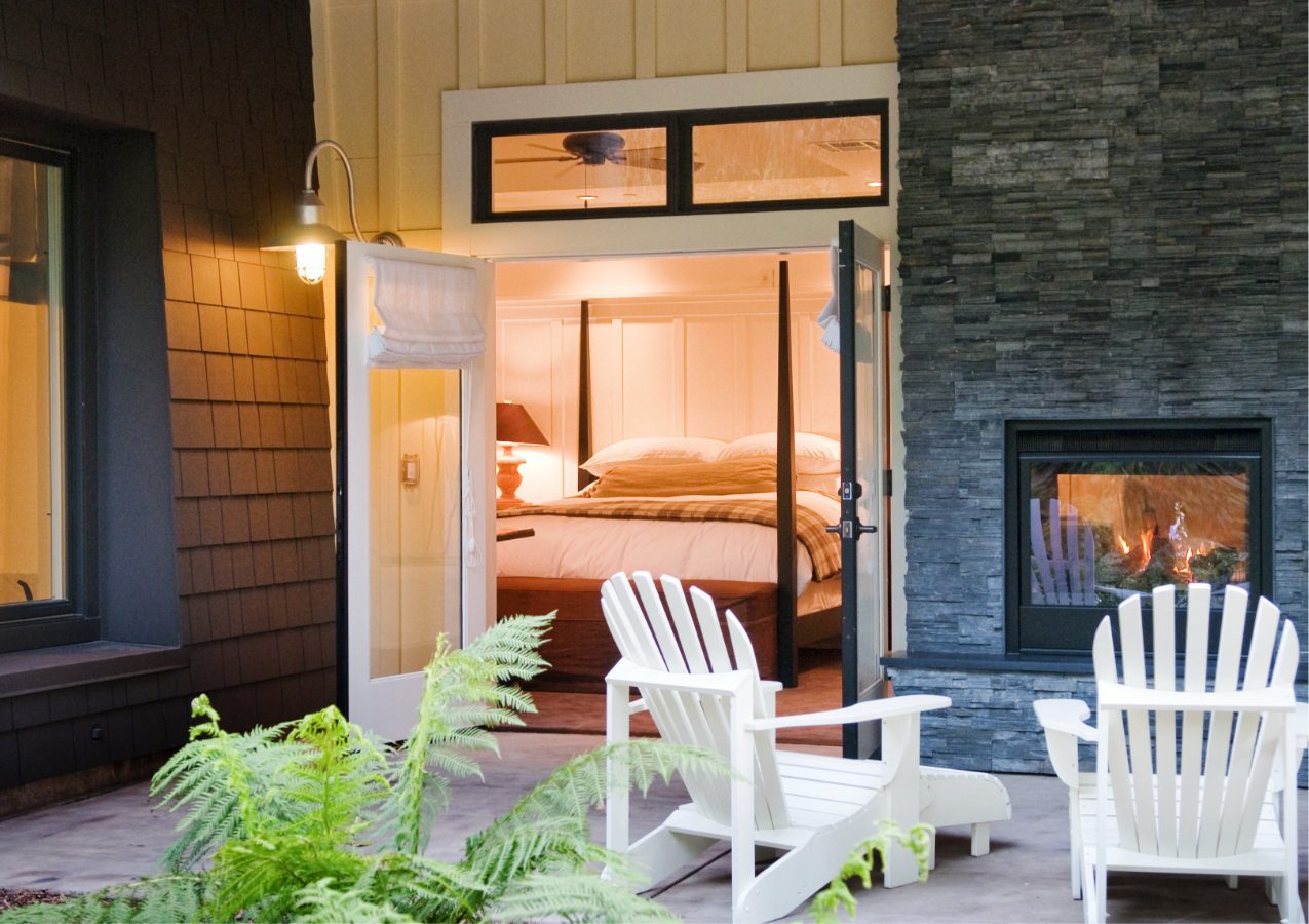 porch view of Barn Junior Suite featuring indoor, outdoor fireplace, white patio wood chairs, and a view inside room with large bed with white bedding